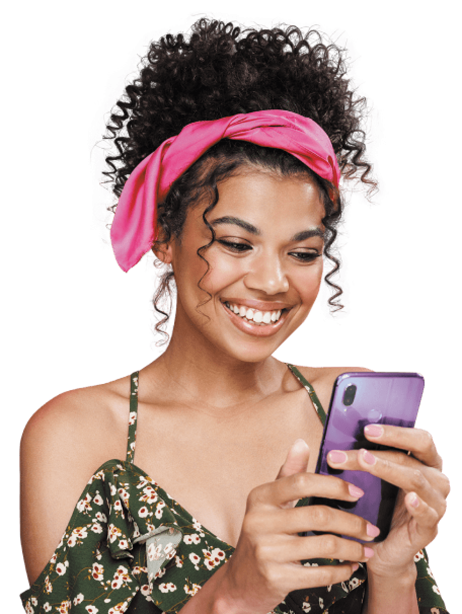Mulher preta, de cabelos cacheados presos com um lenço rosa, sorrindo olhando para o celular que está mexendo na mão.
